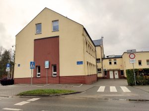 budynek biblioteki w Kolbudach, o kremowej elewacji z elementami czerwonej cegły z białą fugą, w głębi widać parę białych drzwi, na pierwszym planie po prawej znaki drogowe, biały prostokąt z ikoną samochody napis strefa ruchu, białe koło z czerwoną obwolutą, ponize dwa mniejsze, białe prostokątne znaki z nieczytelnymi znakami, po lewej na tle budynku bladoniebieski znak parkingu z literą P, następnie znak przejścia dla pieszych i ponownie znak parkingu z literą P. tło stanowi pochmurne niebo.