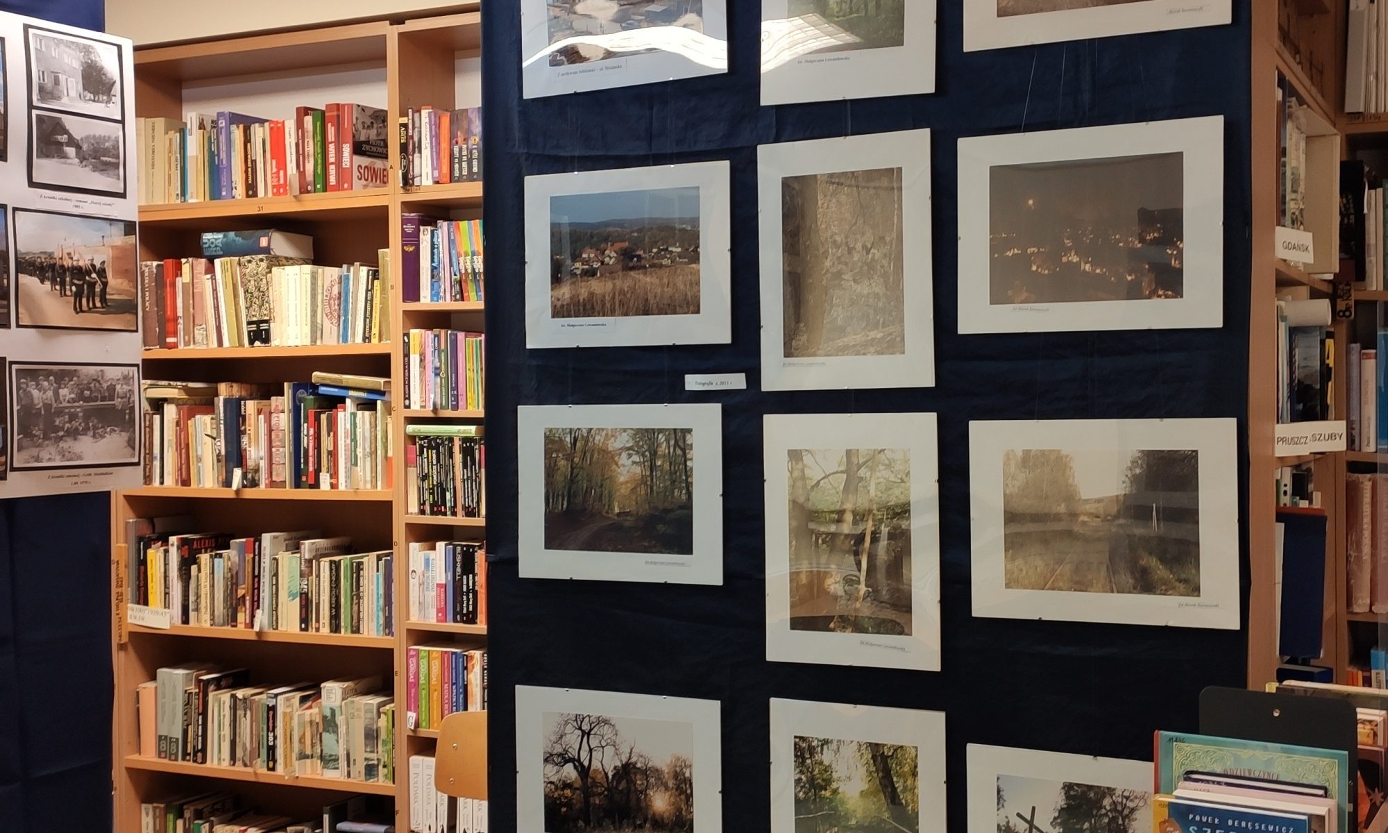 zdjęcie przedstawia wnętrze biblioteki, jasno szara podłoga, kremowe ściany. na pierwszym planie na granatowym tle powieszone są fotografie z białym passe partout, w głębi po lewej widać regały z jasno brązowego drzewa, na których stoją książki