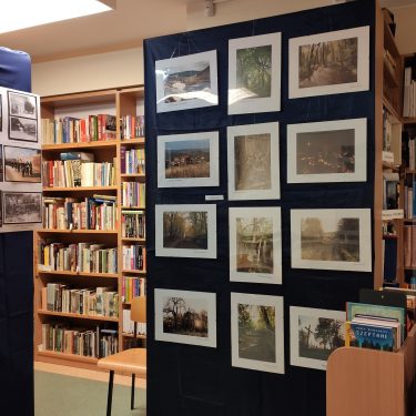zdjęcie przedstawia wnętrze biblioteki, jasno szara podłoga, kremowe ściany. na pierwszym planie na granatowym tle powieszone są fotografie z białym passe partout, w głębi po lewej widać regały z jasno brązowego drzewa, na których stoją książki