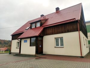 budynek biblioteki w Pręgowie, czerwony dach pokryty dachówką, dwa kominy wypust na dwa okna poniżej wejście, elewacja podbitka z ciemno brązowych desek, poniżej kremowy tynk, po środku drzwi z niebieską tabliczką, parking przed budynkiem z szarej i czerwonej kostki brukowej, szare niebo