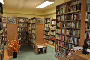 wnętrze biblioteki w Pręgowie, brązowe regały wypełnione książkami, na szczytach powieszone kolorowe obrazy, na podłodze stoi duży czarny wazon z okazałymi żółto pomarańczowymi kwiatami, widoczny jest sufit z białym oświetleniem