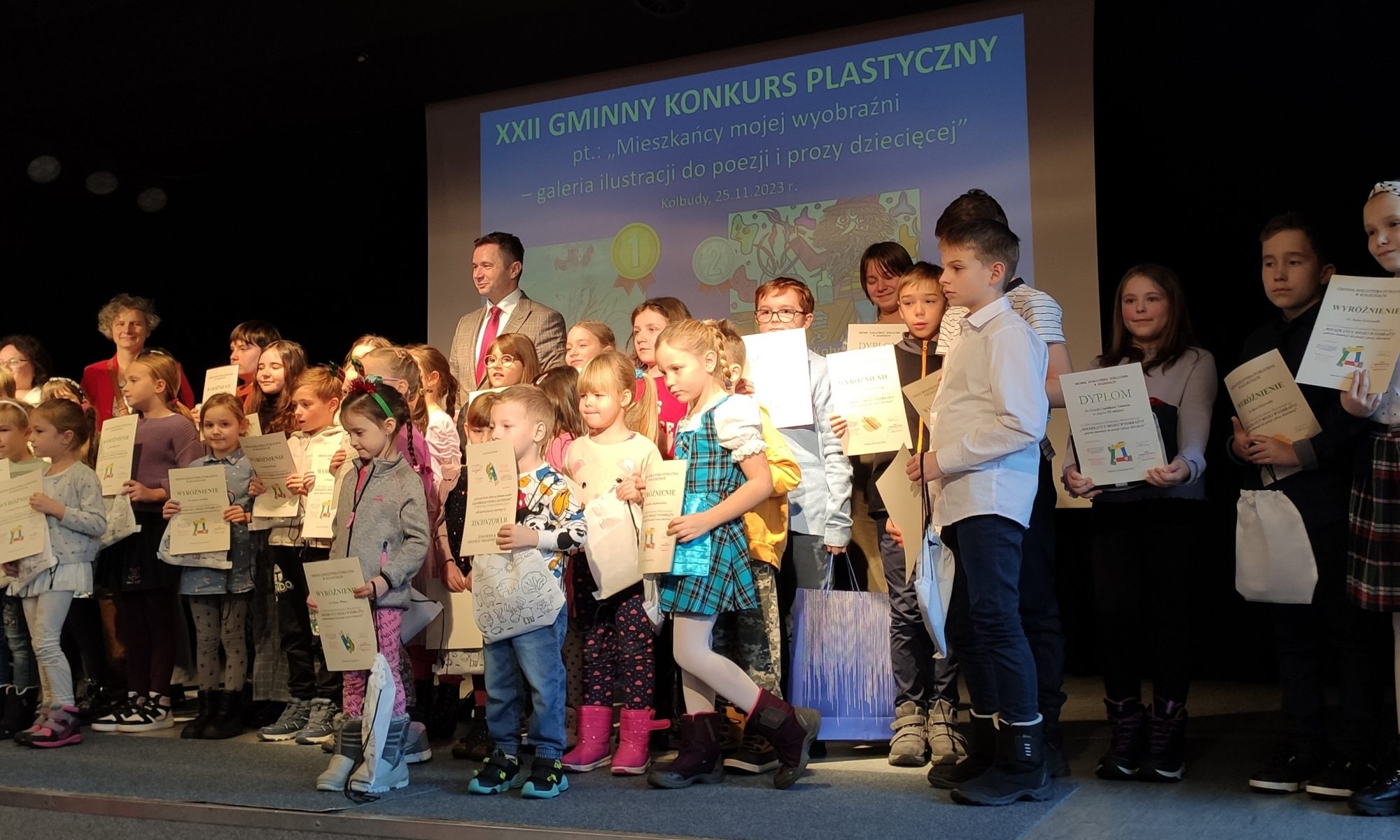 Grupa dzieci i dorosłych stoi na scenie trzymając certyfikaty, uczestnicząc w XXII Gminnym Konkursie Plastycznym. W tle widoczny jest ekran prezentacyjny.
