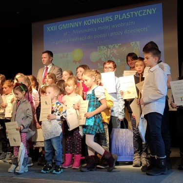 Grupa dzieci i dorosłych stoi na scenie trzymając certyfikaty, uczestnicząc w XXII Gminnym Konkursie Plastycznym. W tle widoczny jest ekran prezentacyjny.