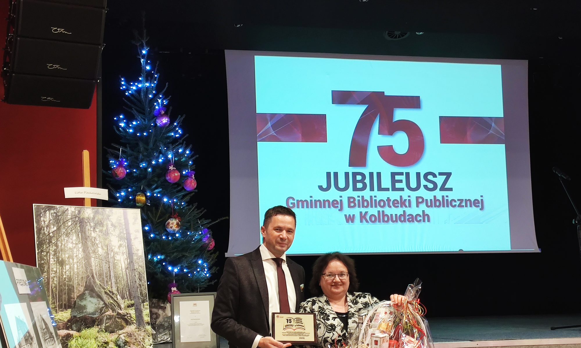 Mężczyzna i kobieta stoją trzymając tabliczkę i ozdobny kosz przed ekranem prezentacyjnym, na którym widnieje napis 75 Jubileusz Gminnej Biblioteki Publicznej w Kolbudach. W pobliżu znajduje się udekorowana choinka i obraz przedstawiający las.