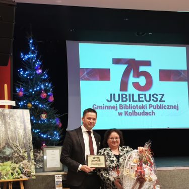 Mężczyzna i kobieta stoją trzymając tabliczkę i ozdobny kosz przed ekranem prezentacyjnym, na którym widnieje napis 75 Jubileusz Gminnej Biblioteki Publicznej w Kolbudach. W pobliżu znajduje się udekorowana choinka i obraz przedstawiający las.