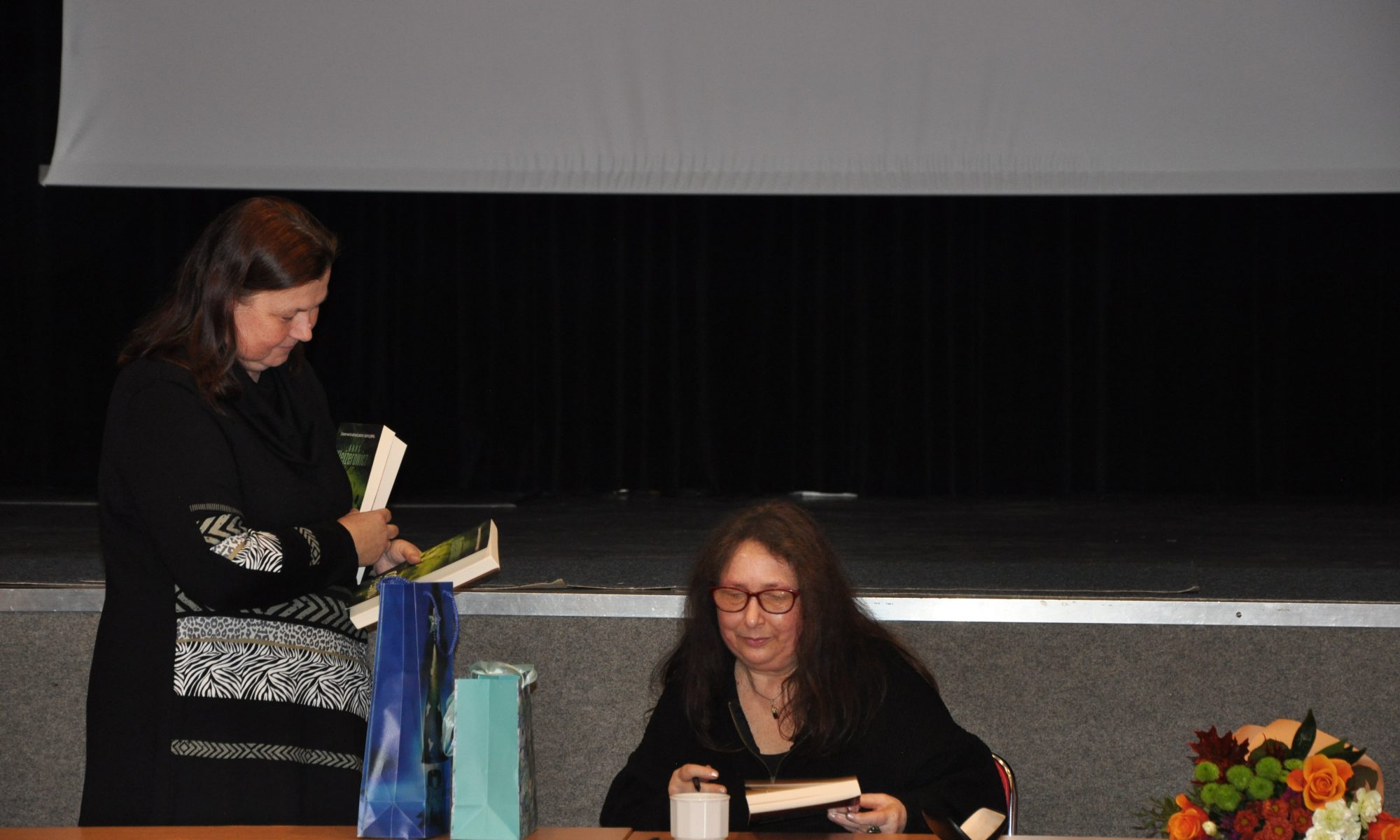 Dwie kobiety z długimi włosami siedzą przy stole. Jedna siedzi i podpisuje książkę, podczas gdy druga stoi i trzyma książkę. Na stole leży niebieska torba z prezentami, a w pobliżu bukiet kwiatów. W tle widać duży ekran.