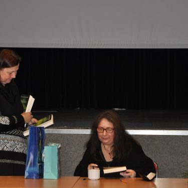 Dwie kobiety z długimi włosami siedzą przy stole. Jedna siedzi i podpisuje książkę, podczas gdy druga stoi i trzyma książkę. Na stole leży niebieska torba z prezentami, a w pobliżu bukiet kwiatów. W tle widać duży ekran.