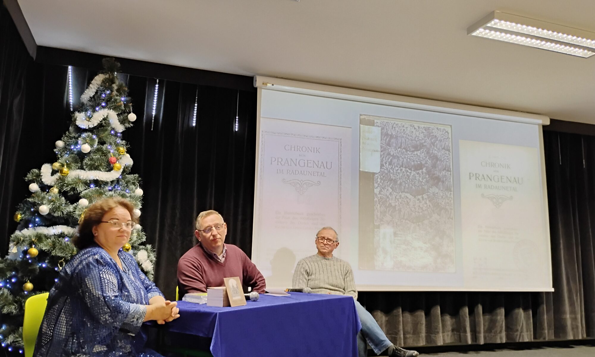 Zdjęcie przedstawia trzy osoby siedzące przy stole. Kobieta siedzi najbardziej po lewej stronie. Za nią jest przystrojona choinka. Tło sceny stanowi ekran ze zdjęciami okładki i strony tytułowej książki Chronik von Prangenau. Zdjęcie okładki jest w środku, a stron tytułowych po prawej i lewej stornie zdjęcia okładki. U góry sali widać sufit z dwiema lampami.