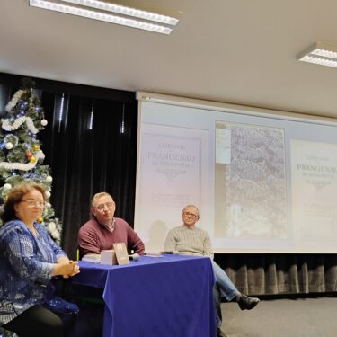 Zdjęcie przedstawia trzy osoby siedzące przy stole. Kobieta siedzi najbardziej po lewej stronie. Za nią jest przystrojona choinka. Tło sceny stanowi ekran ze zdjęciami okładki i strony tytułowej książki Chronik von Prangenau. Zdjęcie okładki jest w środku, a stron tytułowych po prawej i lewej stornie zdjęcia okładki. U góry sali widać sufit z dwiema lampami.