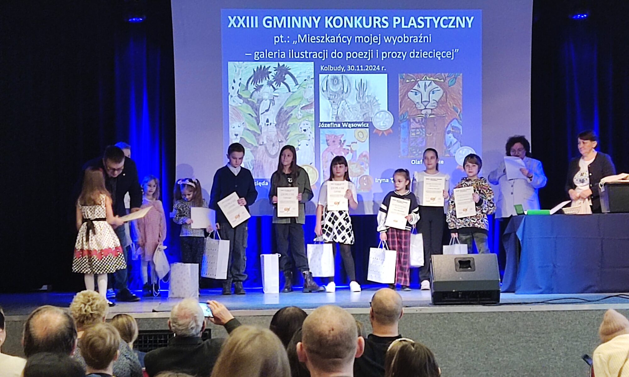 Zdjęcie przedstawia scenę, na której są dzieci z dyplomami oraz troje dorosłych. Kobieta po prawej stronie czyta z kartki, a mężczyzna po lewej stronie wręcza dyplom dziewczynce. Tło sceny stanowi ekran z napisem 'dwudziesty trzeci gminny konkurs plastyczny'. Poniżej napisu wyświetlone są kolorowe prace plastyczne. Przed sceną siedzą ludzie widziani od tyłu.