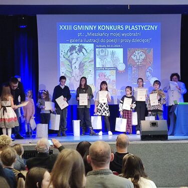 Zdjęcie przedstawia scenę, na której są dzieci z dyplomami oraz troje dorosłych. Kobieta po prawej stronie czyta z kartki, a mężczyzna po lewej stronie wręcza dyplom dziewczynce. Tło sceny stanowi ekran z napisem 'dwudziesty trzeci gminny konkurs plastyczny'. Poniżej napisu wyświetlone są kolorowe prace plastyczne. Przed sceną siedzą ludzie widziani od tyłu.