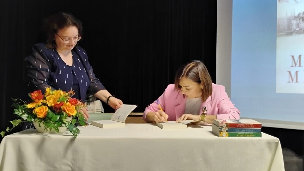 Zdjęcie przedstawia dwie kobiety. kobieta po lewej stronie stoi i trzyma w dłoniach książkę, przed nią jest bukiet kwiatów. Kobieta po prawej stronie siedzi i podpisuje książkę, przed nią wstawione w stos są trzy książki