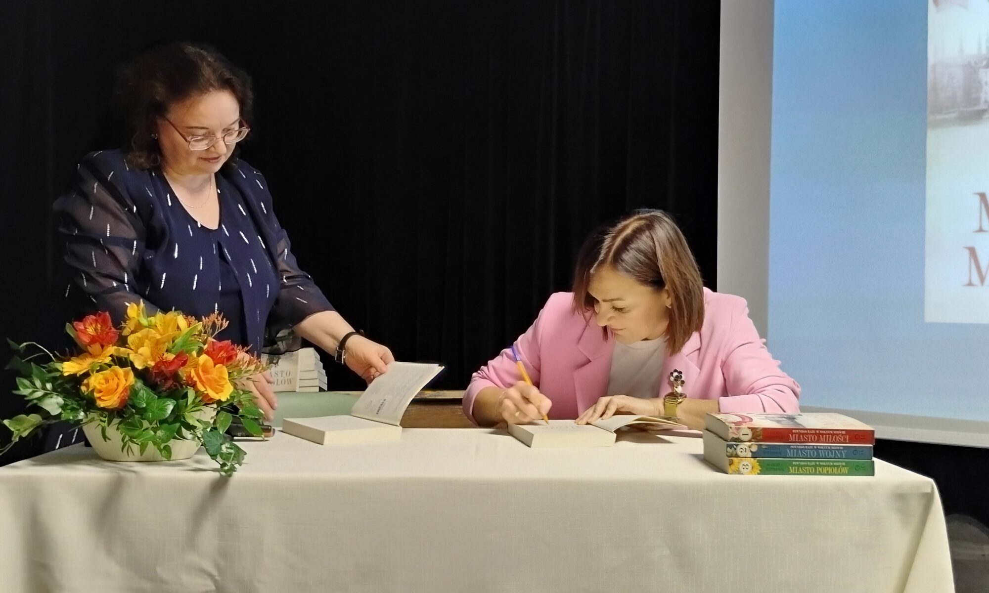 Zdjęcie przedstawia dwie kobiety. kobieta po lewej stronie stoi i trzyma w dłoniach książkę, przed nią jest bukiet kwiatów. Kobieta po prawej stronie siedzi i podpisuje książkę, przed nią wstawione w stos są trzy książki