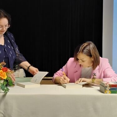 Zdjęcie przedstawia dwie kobiety. kobieta po lewej stronie stoi i trzyma w dłoniach książkę, przed nią jest bukiet kwiatów. Kobieta po prawej stronie siedzi i podpisuje książkę, przed nią wstawione w stos są trzy książki