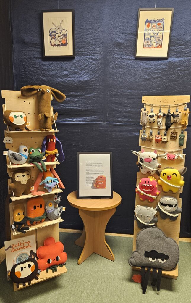 Po obu stronach zdjęcia stoją regały z kolorowymi pluszakami. Nad każdym z regałów wisi obraz. Pomiędzy regałami stoi stoliczek z ramką. Tło zdjęcia jest granatowe