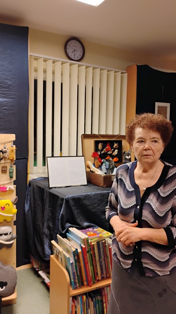 Zdjęcie przedstawia kobietę. Za nią znajduje się otwarta walizka z kolorowymi pluszakami. Poniżej stoją książki z różnokolorowymi okładkami. W górnej środkowej części zdjęcia jest zegar. Po lewej stronie zdjęcia, w połowie ucięty jest regał z kolorowymi pluszakami.