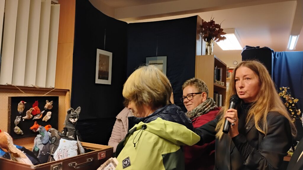 Zdjęcie przedstawia grupę kobiet. Kobieta po prawej stronie, ubrana w czarną kurtkę, trzyma mikrofon. Po lewej stronie zdjęcia znajduje się walizka z kolorowymi pluszakami