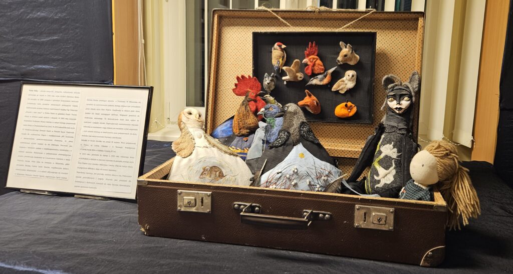 Zdjęcie przedstawia otwartą walizkę, w której znajdują się filcowe pluszaki. Od lewej strony jest sowa, kogut, gołąb i sroka, a po prawej jest laleczka i czarny kot. Za pluszakami są wywieszone filcowe broszki w kształtach różnych zwierząt