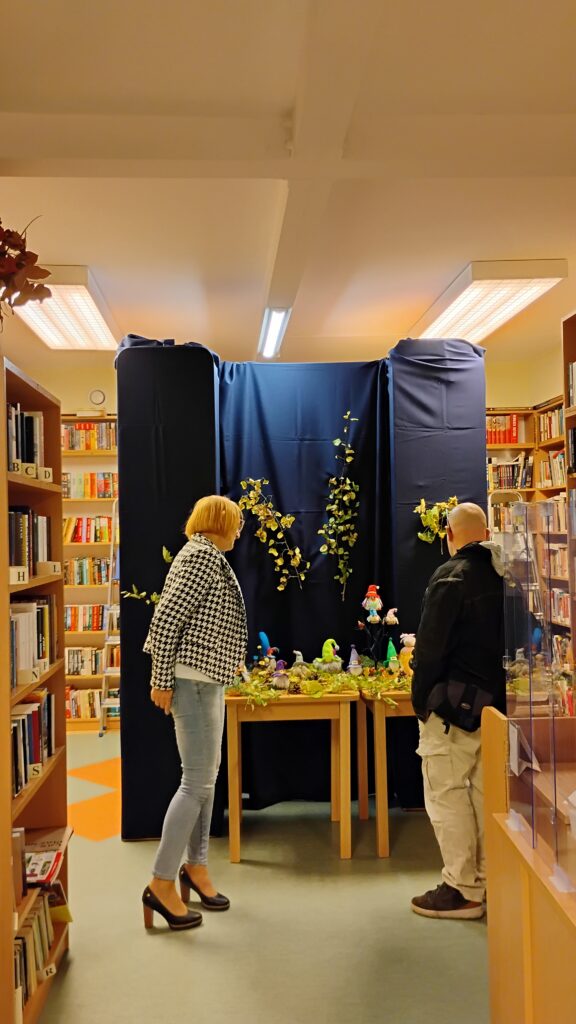 Zdjęcie przedstawia wnętrze biblioteki. W centrum zdjęcia znajduje się stół z kolorowymi skrzatami. Przed nim stoją dwie osoby. Po lewej znajduje się kobieta, po prawej mężczyzna. Obydwoje stoją zwróceni w stronę stołu. Za stolikiem jest granatowe tło z przypiętymi na środku gałązkami z liśćmi