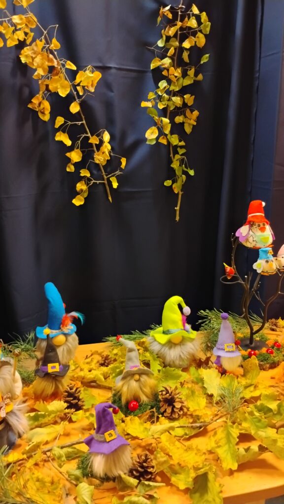 Zdjęcie przedstawia stolik, na którym stoją kolorowe skrzaty ułożone razem z liśćmi i szyszkami. Nad nimi znajdują się dwie gałązki z liśćmi przymocowane do granatowego tła