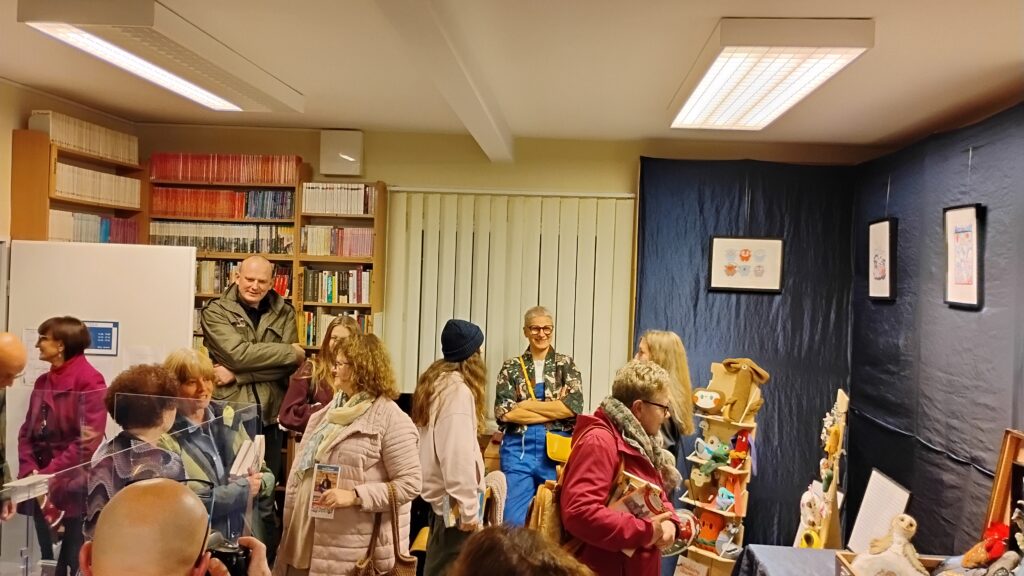 Zdjęcie przedstawia tłum ludzi w bibliotece, Po prawej stronie widać granatowe tłom na którym zawieszono trzy obrazy. Poniżej stoją regały z kolorowymi pluszakami. Po lewej stronie znajdują się białe drzwi, a za nimi regały z książkami