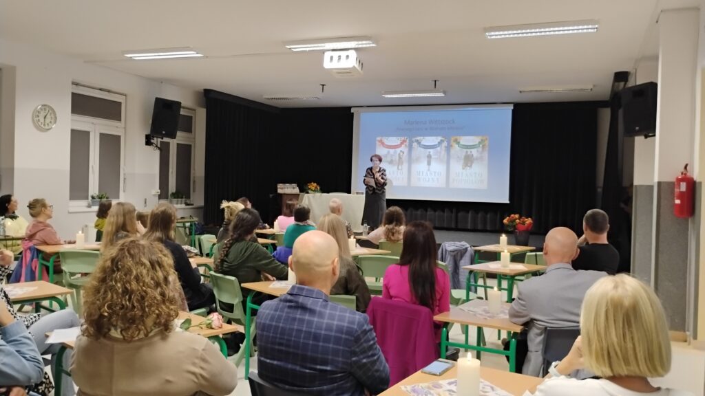 Zdjęcie przedstawia salę wypełniona ludźmi siedzącymi przy stolikach. W dali na scenie stoi kobieta. Znajduje się ona na tle trzech okładek książek. Sufit sali jest biały