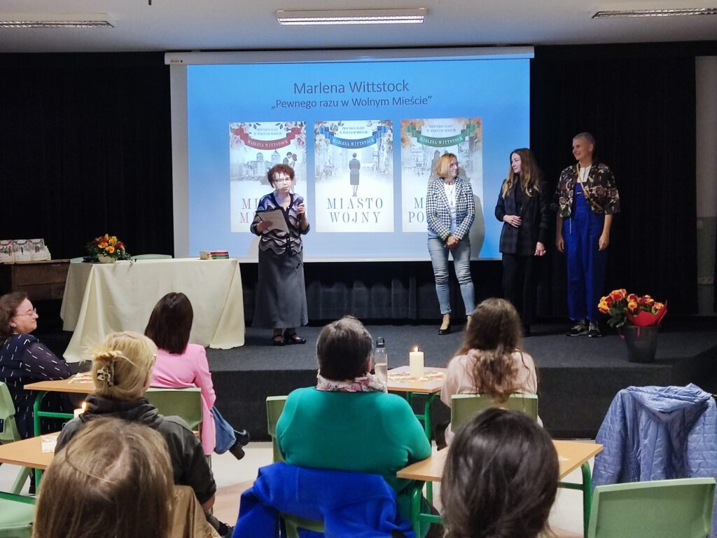 Grupa ludzi siedzi przed sceną. Na scenie po lewej stronie znajduje się stół z białym obrusem, obok niego stoi kobieta. Po prawej stronie sceny znajdują się trzy kobiety, przed nimi jest kosz z kwiatami. Za plecami kobiet jest prezentacja z okładkami książek. U góry prezentacji jest napis "Marlena Wittstock Pewnego razu w Wolnym Mieście"