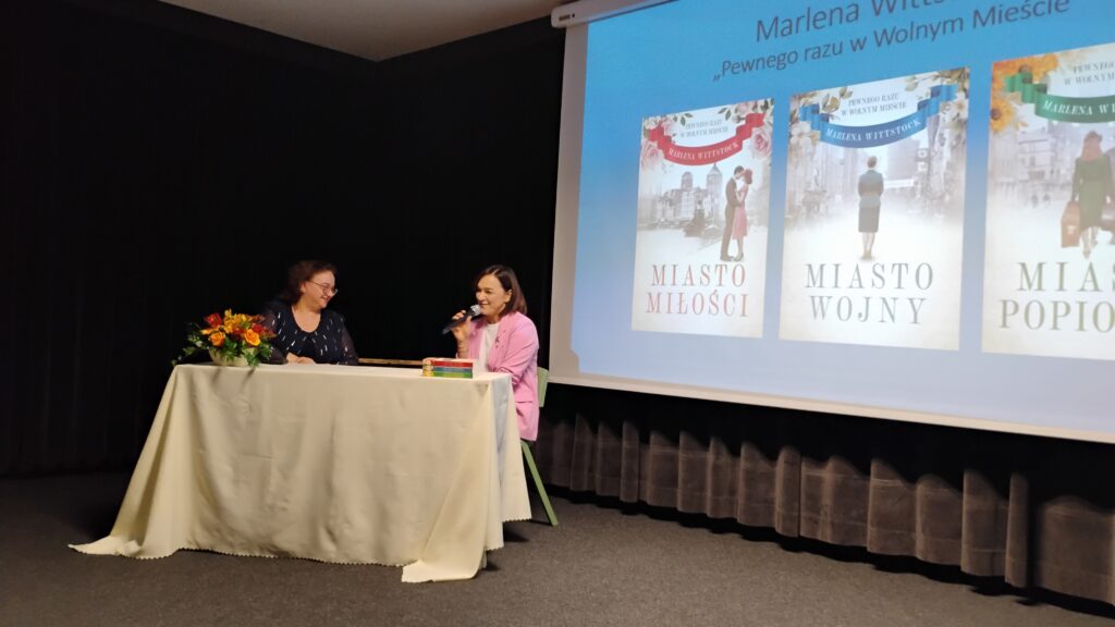 Po prawej stronie zdjęcia jest ekran z prezentacją. Znajdują się na nim trzy okładki książek umieszczone na niebieskim tle. Po lewej stronie zdjęcia znajduje się stół z jasnym obrusem, przy którym siedzą dwie kobiety. Kobieta po prawej stronie ma mikrofon