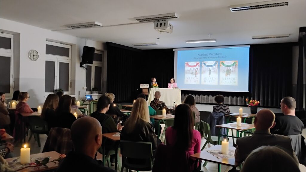 Zdjęcie przedstawia salę, w której odbywa się spotkanie. Dużo osób siedzi zwróconych w kierunku sceny, na której po lewej stronie za jasnym stolikiem siedzą dwie kobiety. Tło sceny stanowi ekran z prezentacją. Przedstawia ona trzy okładki książek na niebieskim tle