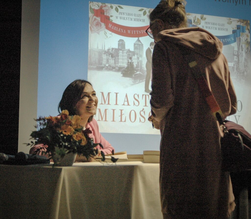 Zdjęcie przedstawia dwie rozmawiające kobiety. Jedna z nich siedzi przy stoliku i jest częściowo zasłonięta przez bukiet kwiatów, który stoi na stoliku. Druga kobieta stoi tyłem i widać jej plecy. Ubrana jest w brązowy płaszcz. Tło zdjęcia stanowi slajd prezentacji z okładką książki