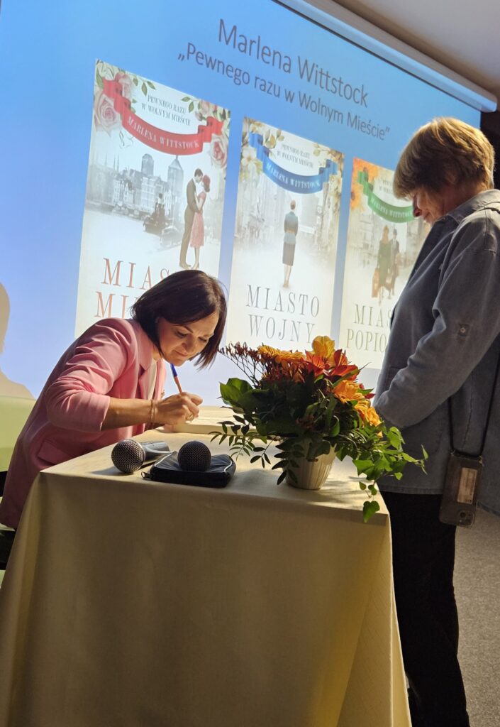 Na zdjęciu znajdują się dwie kobiety widziane z profilu. Stoją one po dwóch stronach stolika. Kobieta po lewej stronie siedzi i podpisuje książkę. Druga kobieta stoi i spogląda w dół obserwując kobietę podpisującą książkę. Pomiędzy kobietami na stole jest bukiet kwiatów. Tło zdjęcia stanowi ekran z trzema okładkami książek