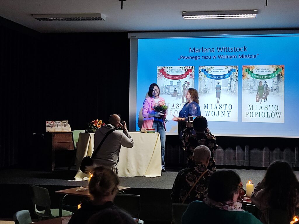 Zdjęcie przedstawia grupę ludzi będących pod sceną. Na scenie znajdują się dwie kobiety. Kobieta po lewej trzyma bukiet kwiatów. Tło sceny stanowi ekran, na którym są wyświetlone trzy okładki książek. Ponad nimi jest napis "Marlena Wittstock pewnego razu w Wolnym Mieście".