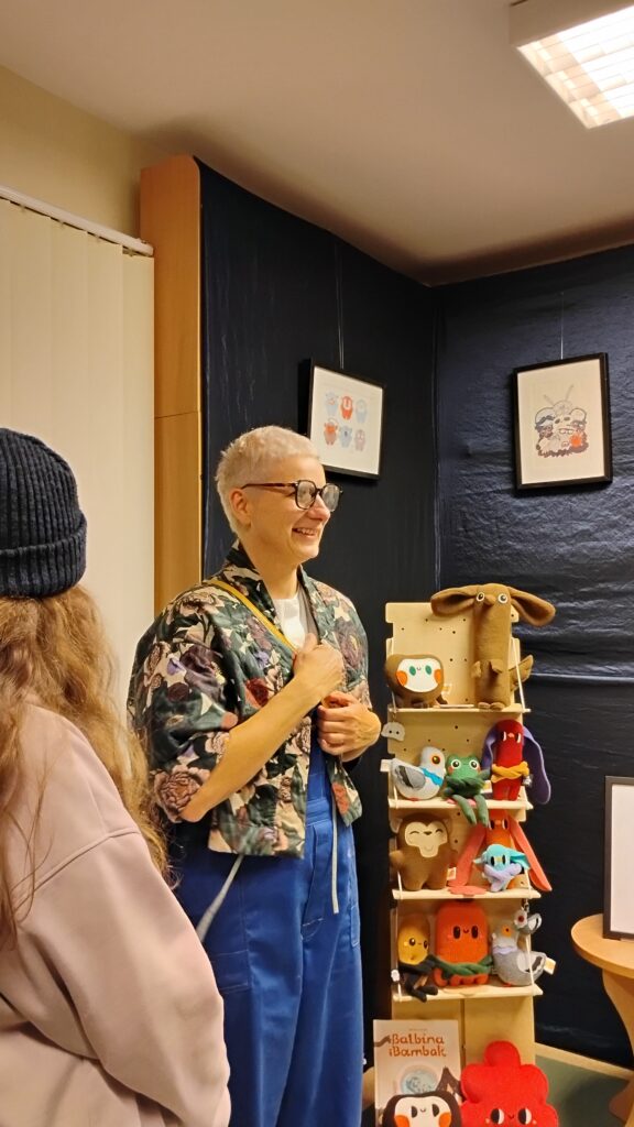 Zdjęcie przedstawia kobietę, która stoi obok regału z kolorowymi pluszakami. Nad nimi są powieszone dwa obrazy na granatowym tle