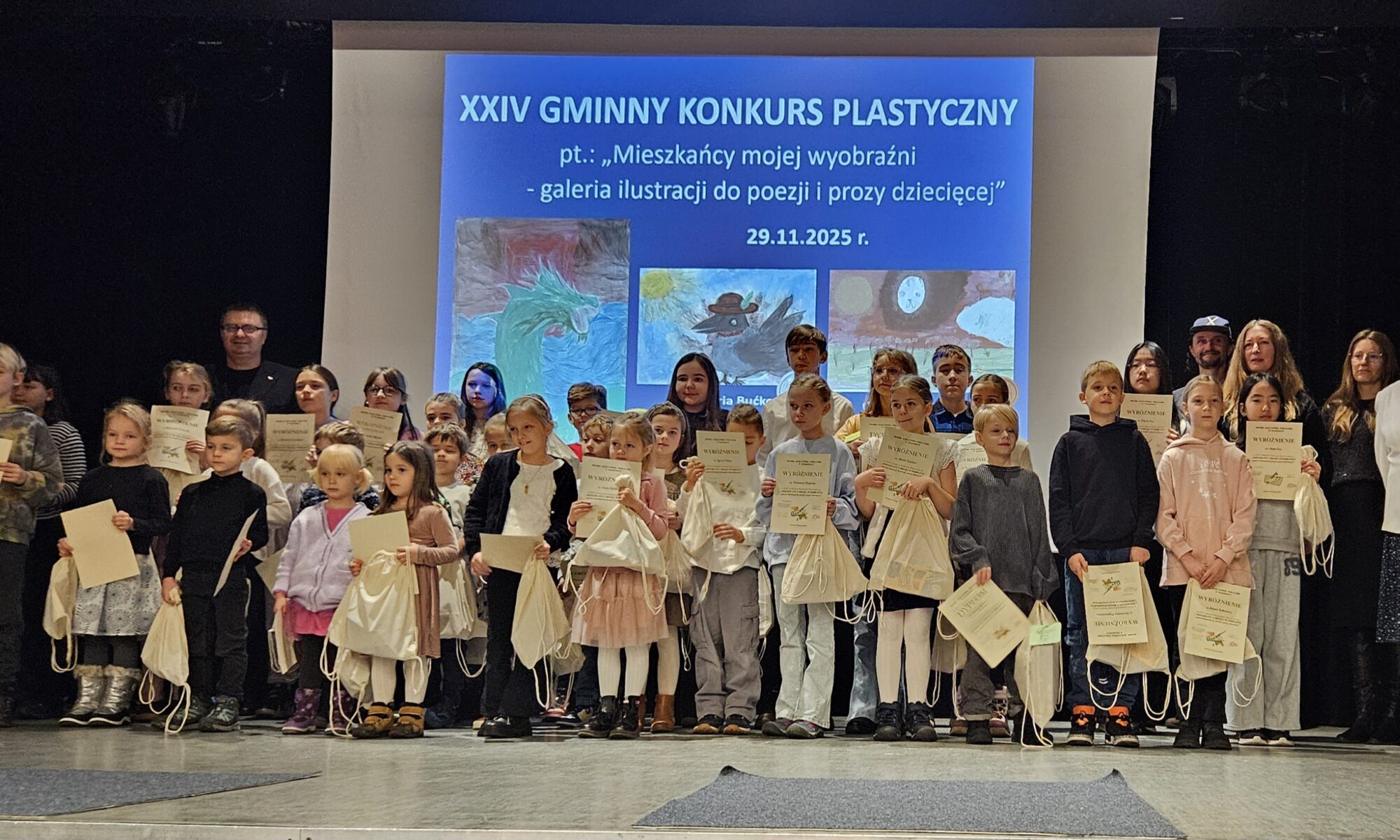 Zdjęcie przedstawia grupę ludzi znajdujących się na scenie. Dzieci trzymają w rękach dyplomy i worki z nagrodami. Za dziećmi znajduje się ekran, na którym jest napis: Dwudziesty czwarty gminny konkurs plastyczny pod tytułem mieszkańcy mojej wyobraźni - galeria ilustracji do poezji i prozy dziecięcej.