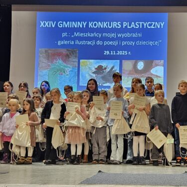 Zdjęcie przedstawia grupę ludzi znajdujących się na scenie. Dzieci trzymają w rękach dyplomy i worki z nagrodami. Za dziećmi znajduje się ekran, na którym jest napis: Dwudziesty czwarty gminny konkurs plastyczny pod tytułem mieszkańcy mojej wyobraźni - galeria ilustracji do poezji i prozy dziecięcej.