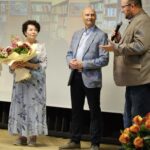 Zdjęcie przedstawia trzy osoby będące na scenie. Po lewej stronie stoi kobieta, która trzyma bukiet kwiatów, pozostałe osoby to mężczyźni. Ten, który jest najbardziej po prawej stronie trzyma mikrofon. Tło stanowi prezentacja z nazwą wydarzenia. Trzydziesty piąty jubileusz gminnej biblioteki publicznej filii w Pręgowie oraz benefis sześćdziesięciolecia pracy zawodowej Pani Barbary Sołczyk