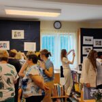 Zdjęcie pokazuje wnętrze biblioteki, w którym znajduje się grupa ludzi. Na przykrytych granatowym materiałem regałach znajdują się zdjęcia stanowiące wystawę fotograficzną. Ludzie przyglądają się wystawie. Na środku sali jest okno, nad nim wisi zegar. Sufit sali jest biały