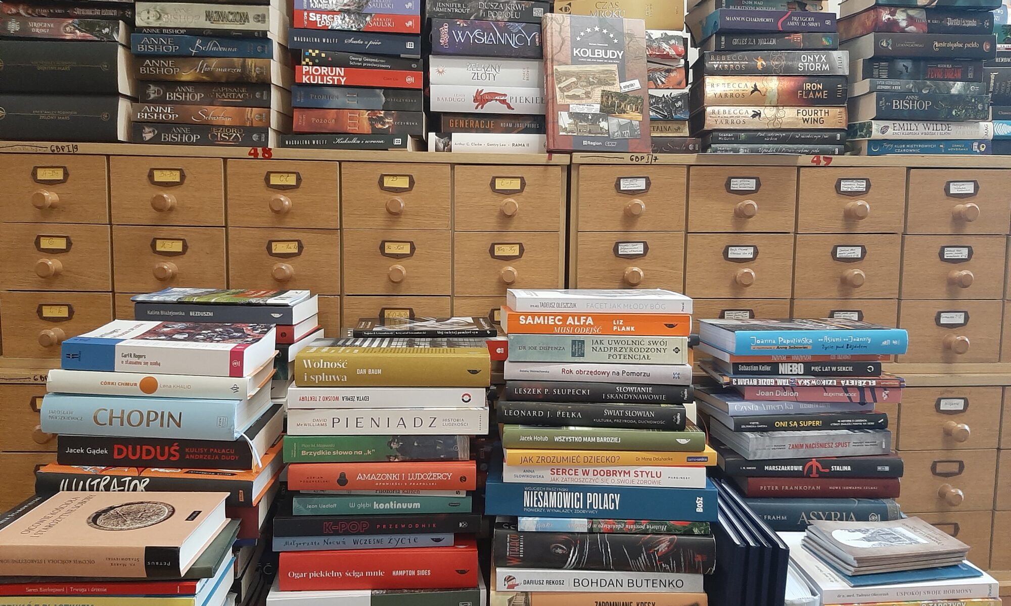 Półka wypełniona książkami, tworzącą wysoką, kolorową stertę z różnorodnymi tytułami.